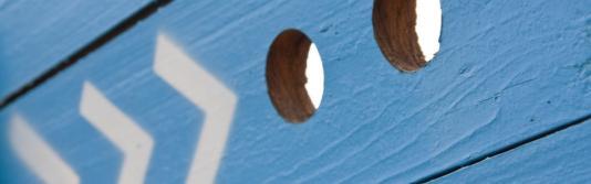 Blaue Holzwand mit Pfeilen, welche auf zwei Gucklöcher weisen