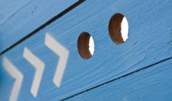 Blaue Holzwand mit Pfeilen, welche auf zwei Gucklöcher weisen